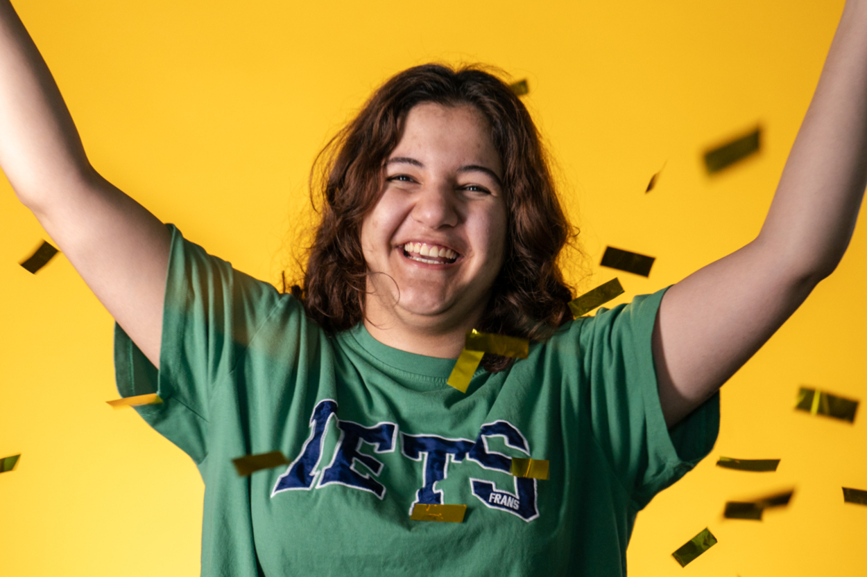 Student celebrating with their arms raised, confetti falling in front of student.