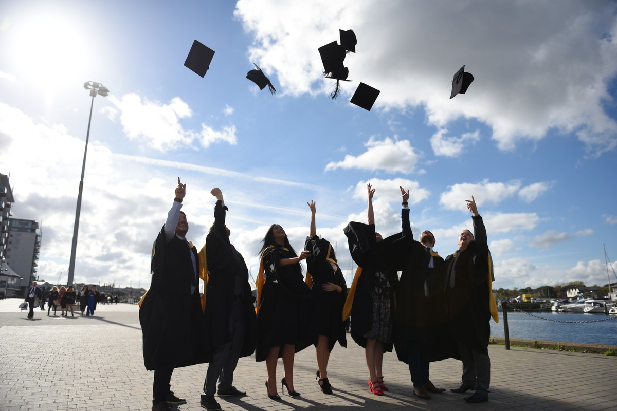 A group of students on Ipswich waterfront dressed in graduation gowns throwing their hats into the air.
